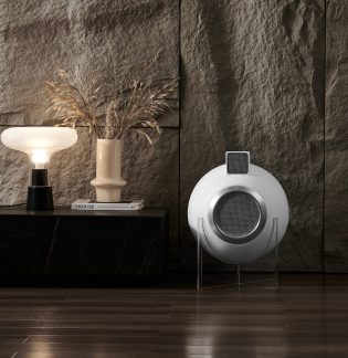 Hvid lampe på sort bord, beige vase med græsser, hvid bog, rund hvid højttaler på gennemsigtig stander, tekstureret væg