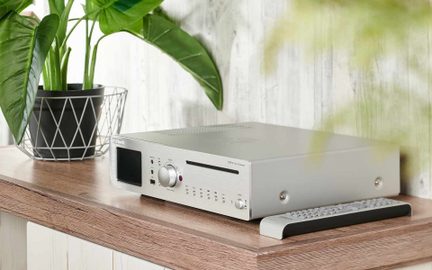 Srebrn ojačevalnik in predvajalnik iz brušenega aluminija, pogled s sprednje strani pod kotom, sprednja plošča z črnim zaslonom, velikim osrednjim gumbom, režo za CD in majhnimi poravnanimi gumbi, postavljen na leseno ploščo