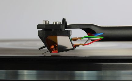 Nærbillede af pladespillerens pickup, sort tonearm og farvede ledninger lagt på en sort vinylplade