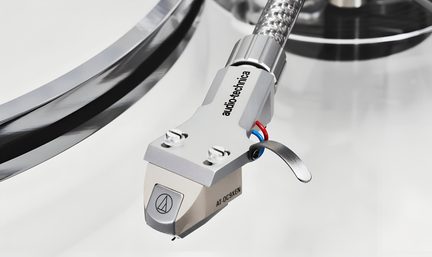 Nærbillede i let fugleperspektiv af en sølvfarvet metal phono-pickup på tonearm med to frontskruer, buet kromhåndtag, synlige røde og blå ledninger samt pickupnål