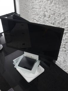 Juoda TV spintelė su plokščia televizoriaus ekranu, pastatyta ant besisukančio balto stovo, kampe prie baltos akmenų sienos