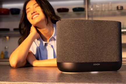 Trådløs sort Denon-højttaler med gråt stof på køkkenbord, smilende kvinde sløret i baggrunden