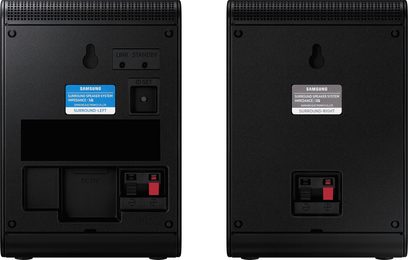 Pár černých matných plastových surround reproduktorů Samsung, pohled zezadu, perforovaná větrací mřížka nahoře, otvory pro montáž na zeď, štítky označující levý a pravý reproduktor, pružinové svorky červeno-černé a konektor napájení DC19V na levém reproduktoru