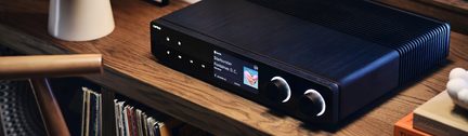 Black hi-fi amplifier with color display and two large knobs, three-quarter view on a wooden shelf