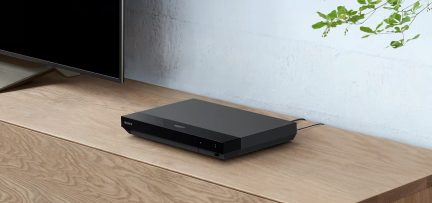 Leitor Blu-ray preto retangular sobre móvel de madeira clara, cabo na parte de trás, tela de televisão à esquerda, parede branca