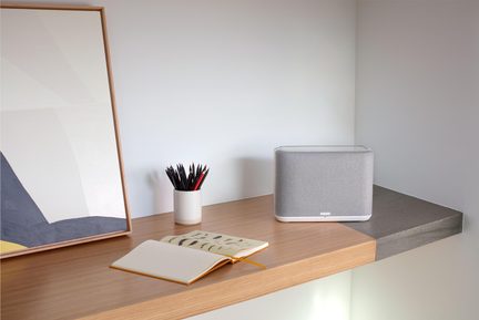 Szara, prostokątna bezprzewodowa głośnik Bluetooth na drewnianym biurku, obok otwartego notesu i kubka z ołówkami