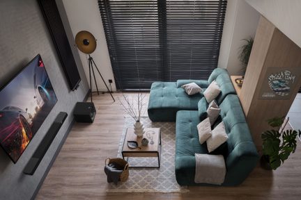 Turkis hjørnesofa, rektangulært salongbord, teppe med geometriske mønstre, svarte venetsianske persienner, grønne potteplanter