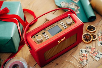 Rød bærbar radio i kunstlæder, set i en let hældende trekvartsvinkel, med matchende håndtag, gyldent top-panel med integreret rektangulært display, to store cremefarvede runde knapper, seks hvide rektangulære knapper og gyldent metalhøjttalergitter