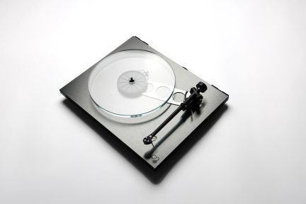 Toca-discos visto de cima, prato circular em vidro transparente, braço de leitura preto, base retangular com acabamento metálico