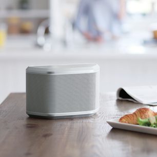 Yamaha WX-030 white wireless connected speaker, front view, placed on a wooden table next to a plate with a croissant