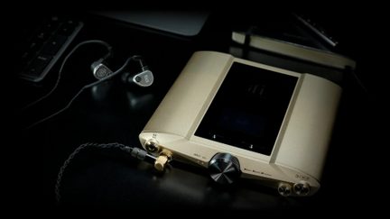 Zlatý audio přístroj s černým displejem, otočným tlačítkem, jack konektory a černými sluchátky připojenými krouceným kabelem