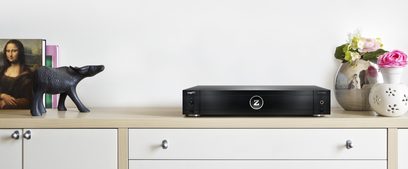 Síťový audio přehrávač Zappiti Pro 4K HDR černý, pohled zepředu, umístěný na světlém dřevěném nábytku mezi soškou hrocha a vázou s květinami