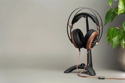 Svart og tre hodetelefoner på svart stativ, kobberfarget spiralformet kabel, grønn plante i potte til høyre, grå bakgrunn