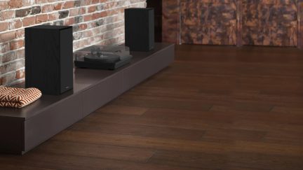 Dvě černé dřevěné reproduktory a černý gramofon s průhledným krytem na nízkém hnědém nábytku, šikmý pohled