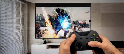 Proyector Optoma UHD35 proyectando una imagen de primer plano de un personaje de videojuego con efectos luminosos y explosivos, sostenido en una mano que sujeta un mando de Xbox