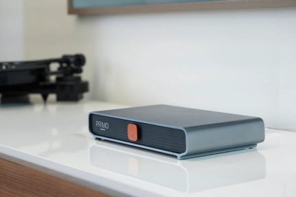 Rektangulært gråt kabinet, sort front med orange knap i midten, placeret på en hvid reflekterende overflade