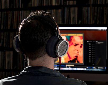 Un bărbat văzut din spate purtând căști audio în fața unui ecran care afișează o imagine colorată și o listă de titluri muzicale
