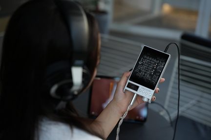 Fehér Questyle audiofil zenelejátszó fekete kijelzővel, nagy fül köré simuló fejhallgatóval az használó nő fején