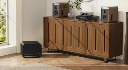 Ruskea puinen taso, jossa on ovaalisti uritetut ovet, kaksi puista ja mustaa elektroniikkaa sisältävää kaiutinta päällä, musta subwoofer lattialla