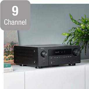 Denon AVC-X3700H sort hjemmebiografsforstærker, set forfra, placeret på hvidt møbel, grøn plante til højre, "Channel 9"-skilt til venstre