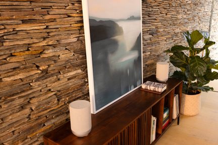 Paar zylindrischer, beiger Stoff-Lautsprecher auf einem TV-Möbel aus Holz vor einer Steinwand, mit Bild und grüner Pflanze