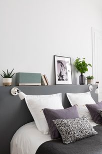 Værelse med grå væg, hvid reol, planter, rammer, bøger, lamper og seng med hvide og lilla puder