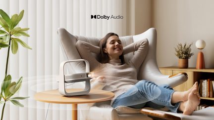 White portable video projector with Dolby Audio, modern square design on coffee table, cozy living room with relaxed woman