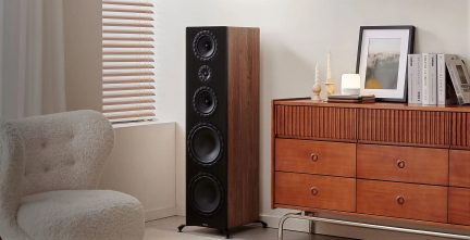 Ahşap kaplamalı ve siyah ön yüzlü hi-fi kule hoparlör, ahşap bir konsolun ve stor perdeli bir pencerenin yakınındaki bir salonda