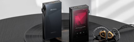 Du juodi nešiojami garso grotuvai su jutikliniu ekranu ir šoniniais mygtukais, kartu su auksinėmis ausinėmis ant vinilo plokštelės.