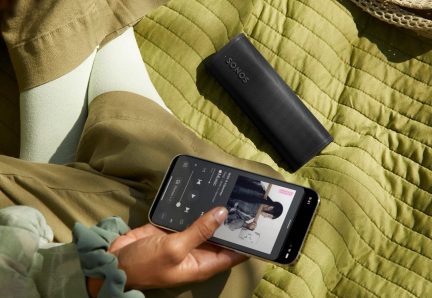 Altoparlante portatile nero Sonos su coperta verde, mano che tiene smartphone con applicazione musicale visualizzata
