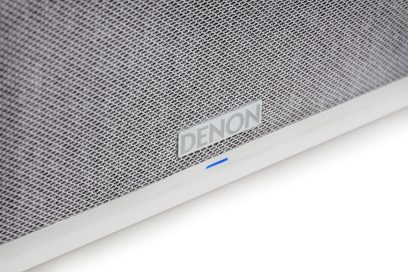 Biała inteligentna kolumna Denon Home 250, zbliżenie na front z szarej tkaniny z metalowym logo i niebieską diodą LED
