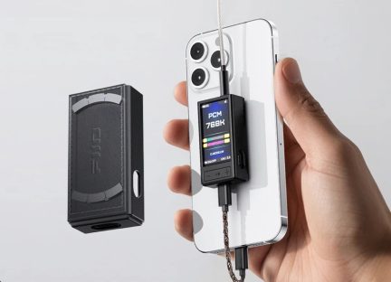 DAC/amplificador de áudio preto em metal com tela colorida e botões +/-, conectado por cabo a um smartphone branco, estojo preto ao lado