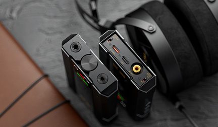 To sorte, bærbare lydkabinetter i metal set let skråt ovenfra, med affasede kanter, en centreret rillet volumenkontrol på den ene, forskellige hovedtelefonudgange, USB-C-port, gul koaksialindgang og farvede LED-indikatorer