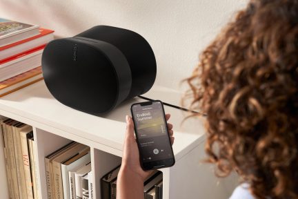 Front view of a black Sonos smart speaker, controlled by a hand holding a smartphone displaying the Sonos app