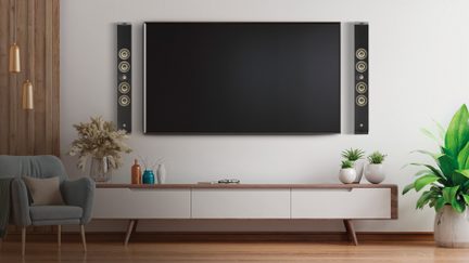 Stort fladskærms-tv i sort med smal metalramme og glasoverflade, set forfra, monteret på væggen mellem to sorte højtalerkolonner med blank facade, hver med fem runde højttalere
