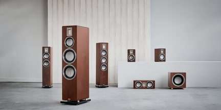 Sæt af rektangulære højttalere i brun træfinér, grå runde højttalere med sort kant og rektangulære diskanter opstillet lodret, set forfra i et minimalistisk interiør