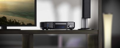 Denon S-102 Blu-ray-afspiller i sort set forfra, blå LCD-skærm, på træhylde med udsigt over strand i baggrunden