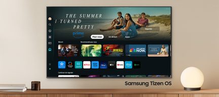 Televizor Samsung s operačním systémem Tizen zobrazující uživatelské rozhraní s aplikacemi pro streamování, umístěný na světlém dřevěném nábytku s bílou kulatou lampou a knihami