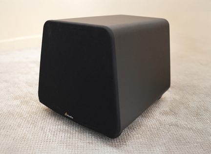 Caixa de som preta, formato trapezoidal, cantos arredondados, logotipo dourado visível na parte inferior, colocada sobre um carpete bege