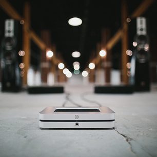 BlueSound NODE X srebrny odtwarzacz audio sieciowy, widok z przodu na betonowej powierzchni, rozmyte tło z butelkami alkoholu
