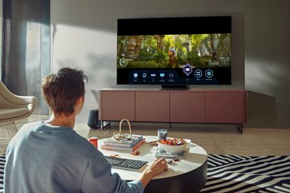 Televizor Samsung QE85QN900A zobrazující les, před osobou sedící zády na růžové pohovce v obývacím pokoji