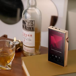 Lettore audiophile Astell&Kern SP3000T in rame in piedi, schermo acceso, su libro dorato, bottiglia e bicchiere a sinistra, candela accesa