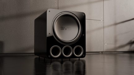 Sort subwoofer i sort, set forfra, med 3 cirkulære porte nederst, SVS-logo i midten, gråt gulv og væg i baggrunden