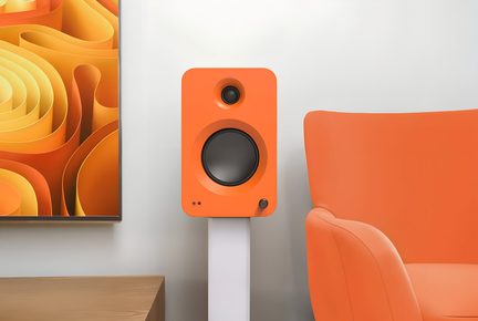 Kompakter, orangefarbener Lautsprecher in mattem Kunststoffgehäuse, Frontansicht, mit eingelassenem schwarzem Hochtöner oben, dunkelgrauem, rundem Tieftöner in der Mitte, zwei kleinen LEDs unten links und schwarzem Drehregler unten rechts, auf einem weißen Standfuß platziert