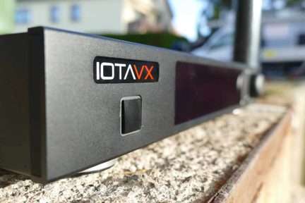 Schwarzer IOTAVX-Verstärker in schräger Vorderansicht, mit zentralem Knopf und großem dunklem Display an der Frontseite