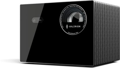 Czarny projektor Valerion VisionMaster Max, front z błyszczącego szkła z dużym, okrągłym obiektywem otoczonym napisami oraz małym, prostokątnym czujnikiem, boczna obudowa z szarego, żebrowanego metalu, widok z prawego przedniego trzech czwartych.