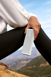 Sonos Roam Lunar White Bluetooth Speaker held in a hand against a mountainous landscape background