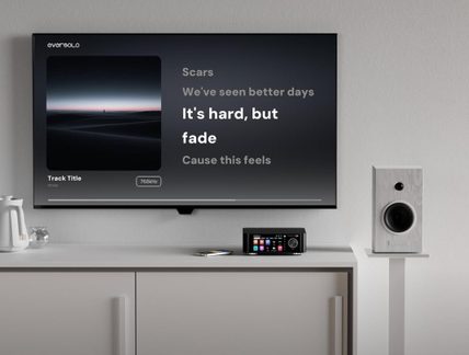 Black network audio player with color display on white cabinet, with wall-mounted TV and white hi-fi speaker