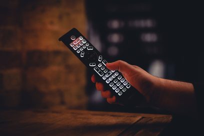 Télécommande iR14 Cyrus tenue par une main, vue de côté, boutons rétroéclairés, fond flou sombre, surface en bois en bas