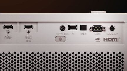 Vedere din spate în prim plan a unui videoproiector din plastic mat alb, echipat cu două porturi HDMI, un port USB, un conector optic SPDIF, o priză serială RS-232, prize audio de 12 V și jack de 3,5 mm, precum și o grilă largă de ventilație perforată.